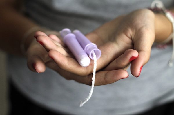 A woman with red-painted nails holds her palms out in front of her, on which, lies a light purple-colored tampon.