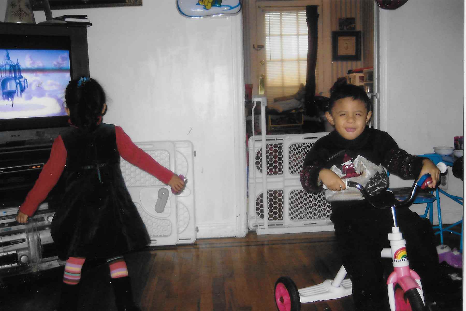 A little girl stands in front of a TV so her back is facing the camera. Her twin brother is looking at the camera with a scrunched face while riding a tricyle inside their family's living room.